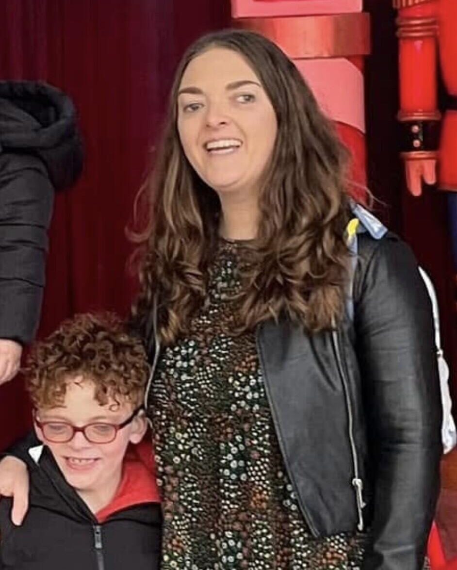 A boy and a woman standing closely together, both smiling. The woman has medium-length, wavy brown hair, and is dressed in a black leather jacket over a knee-length floral dress. The boy, with short curly hair and red glasses, is in a black puffer jacket topped with a red hoodie. They are positioned in front of a rich red backdrop, with soft spotlighting creating a warm ambience. The composition includes a slight angle, emphasising their expressions and the festive context.