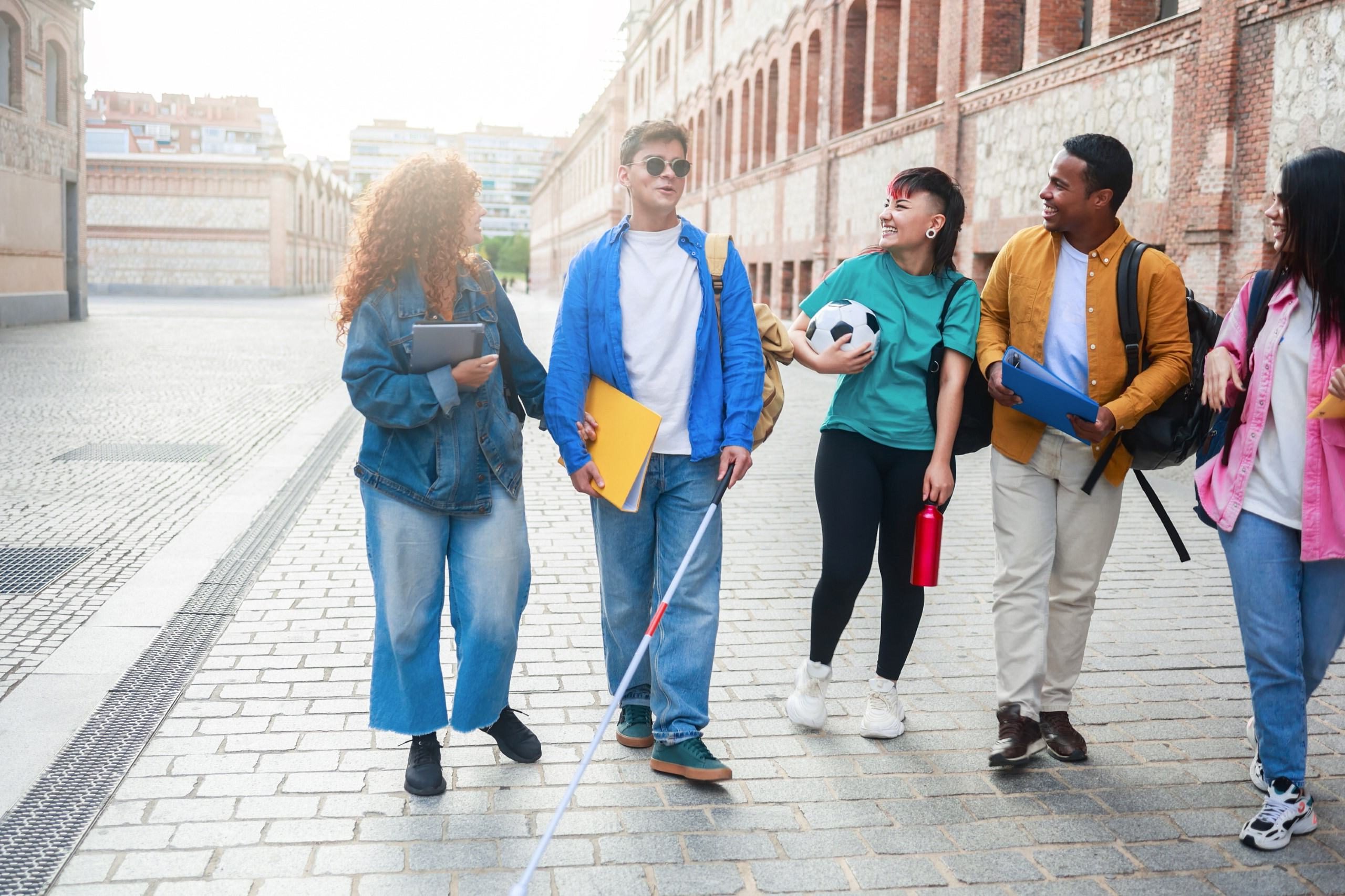 The picture features five young individuals walking on a textured cobblestone pathway with a brick and stone backdrop. Two of them are positioned on the right, while three occupy the left. On the centre right is a young man using a white cane. He has short brown hair and is dressed in a blue jacket and jeans. He links arms with a curly ginger-haired young woman wearing all denim. On the centre left stands a young woman in a green shirt and jeans holding a football, a young man holding a binder and another young woman wearing a pink shirt. They are all smiling and looking at the young man with the cane as if he had just said a joke, creating a dynamic composition. The lighting is bright, suggesting it's a sunny day, adding a lively atmosphere to the scene.