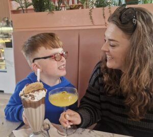 A young boy with short ginger hair and red glasses expresses joy as he holds a tall glass of chocolate milkshake with whipped cream and a cookie. To the right, a woman with long, curly brown hair and a black and grey striped sweater smiles back at him while holding a yellow cocktail in a stemmed glass. The table is set with silver utensils and a marble surface, and there are decorative plants in the background, contributing to a warm and inviting café atmosphere.