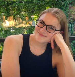 A photo of a young women, Joné sitting at a table wearing black glasses smiling at the camera. She is leaning her head on left fist. She has light brown hair and is wearing a black sleeveless high neck top.