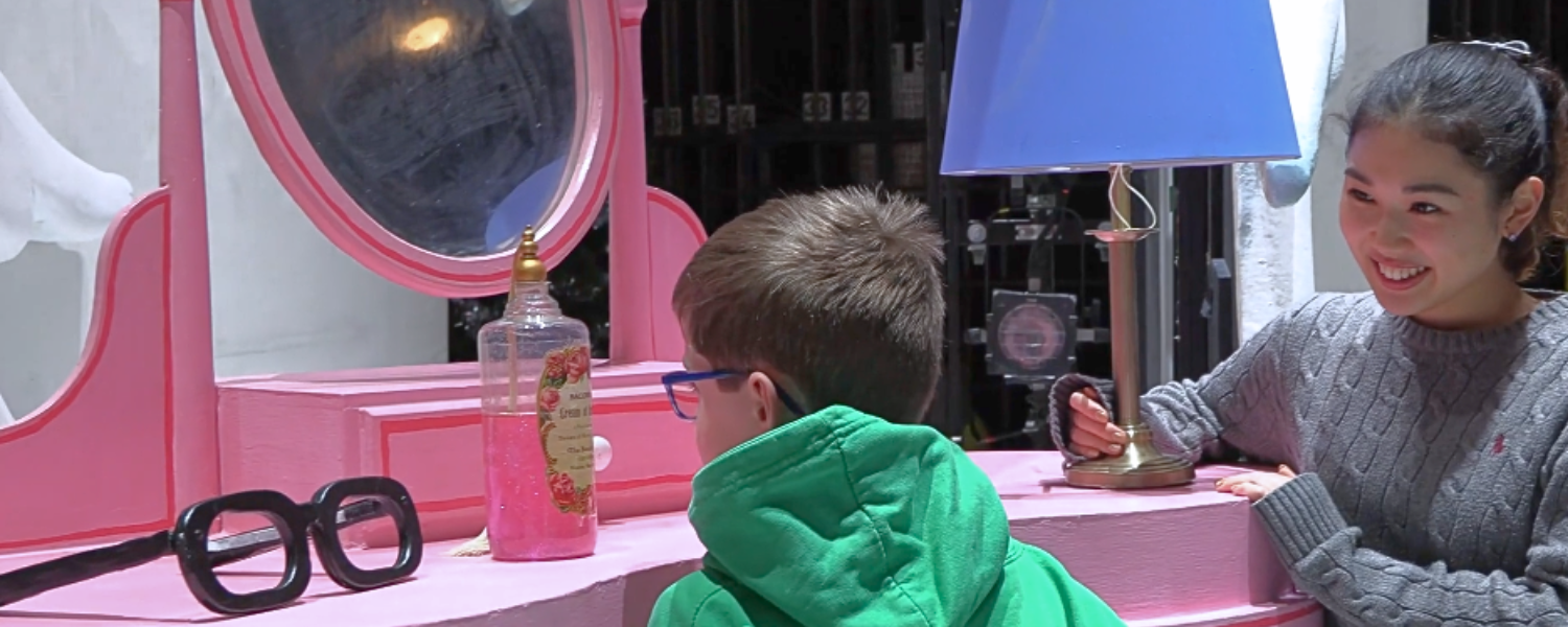 A small boy, on the stage of The Snowman, exploring the set. He's standing in front of an over-sized pink dressing table. Next to him is a staff member from the theatre, talking him through the set and props.