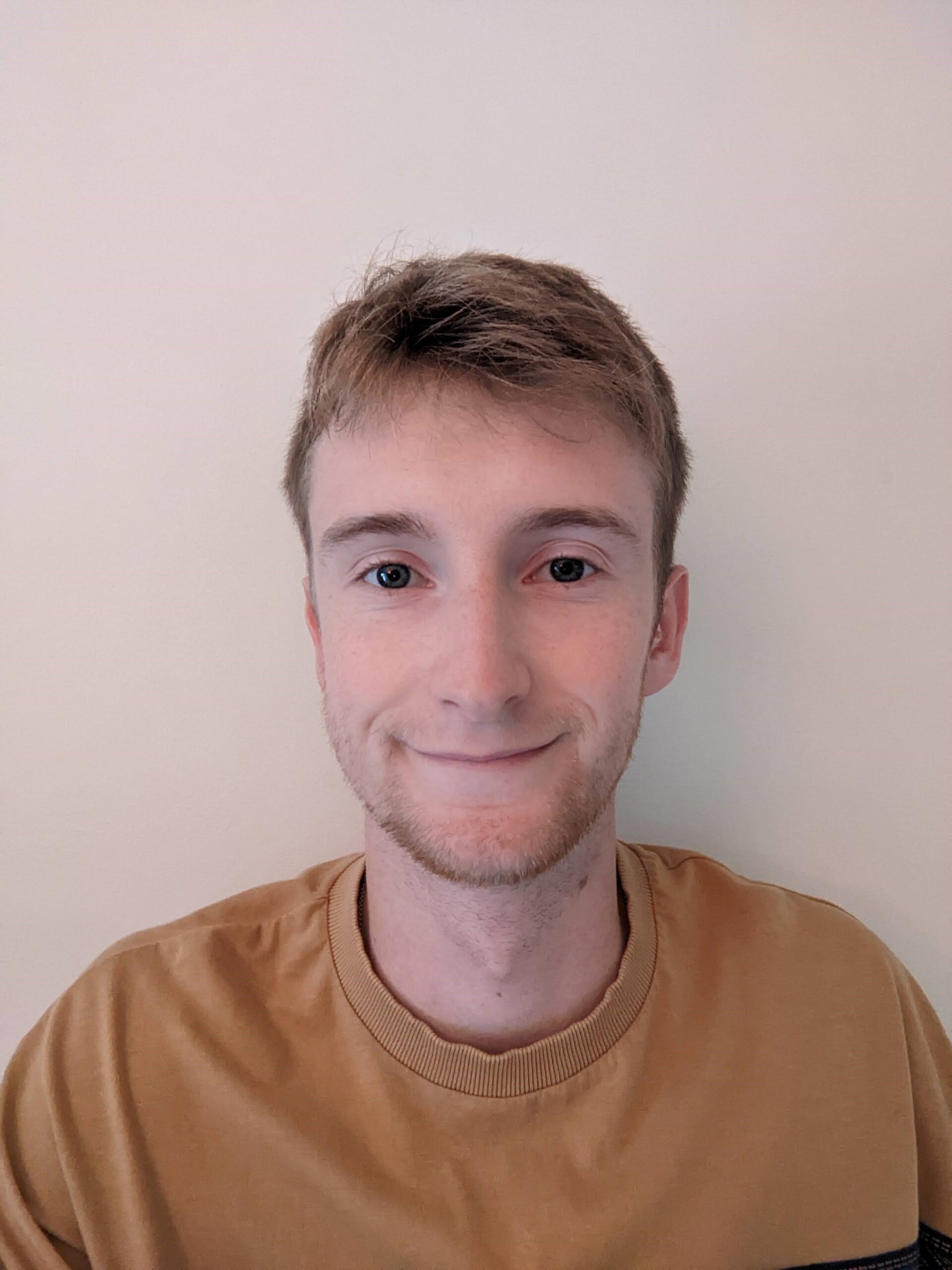 A light-skinned male with blonde hair, and a light beard wearing a beige t-shirt smiling at the camera.