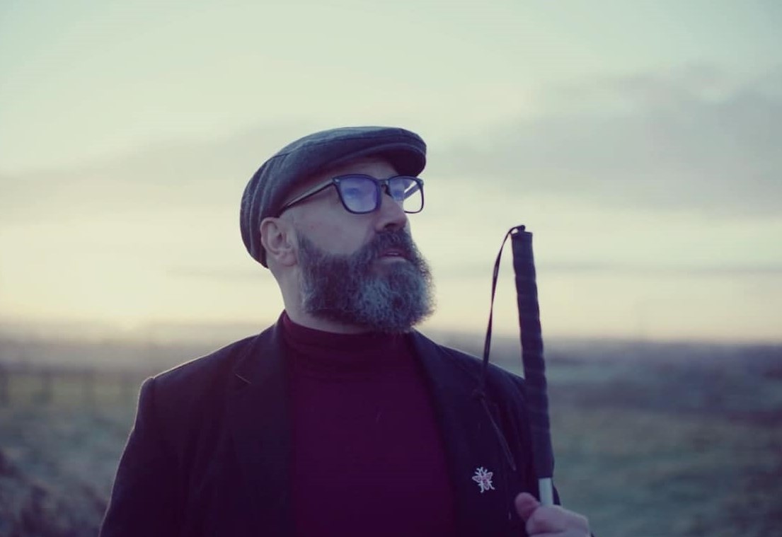 A photograph of Dave The Blind Poet. He is standing in a field wearing a black coat, burgundy roll-neck top and a grey hat. The top of his cane is in view on his left. His head is faced slightly to the left and tilting upwards.