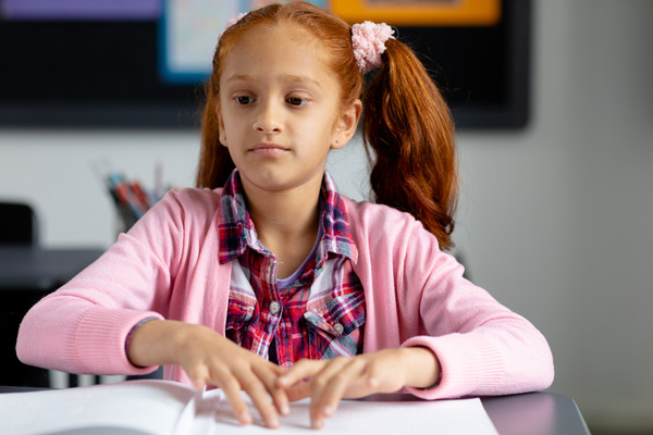 A young girl with red hair in pigtails sits at a desk, intently reading a book written in Braille. She is wearing a pink cardigan over a red and blue plaid shirt. Her fingers are carefully tracing the raised dots.