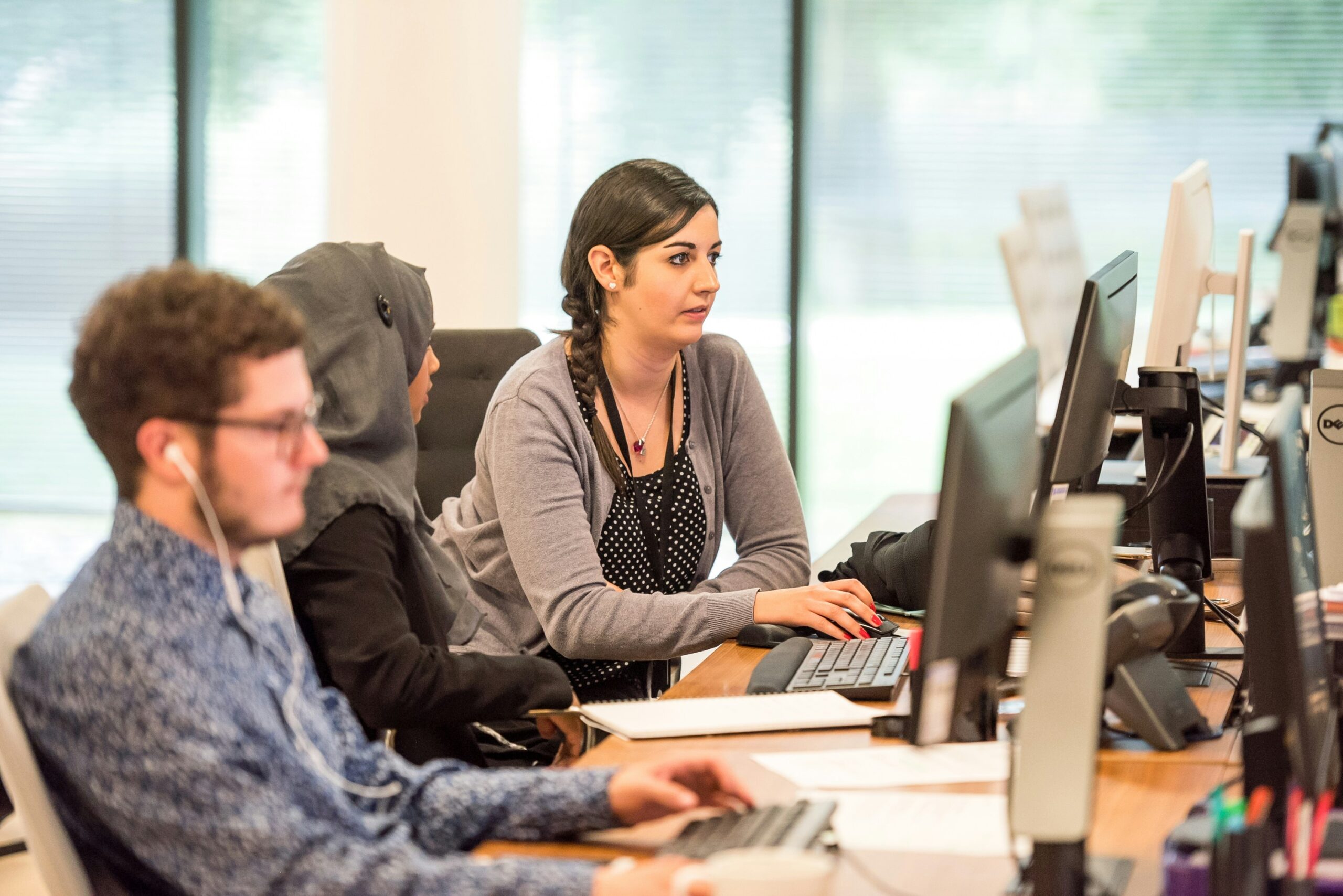 Two women sitting at a computer in an office, one is helping the other with work. There is a man sitting at his computer to their right.